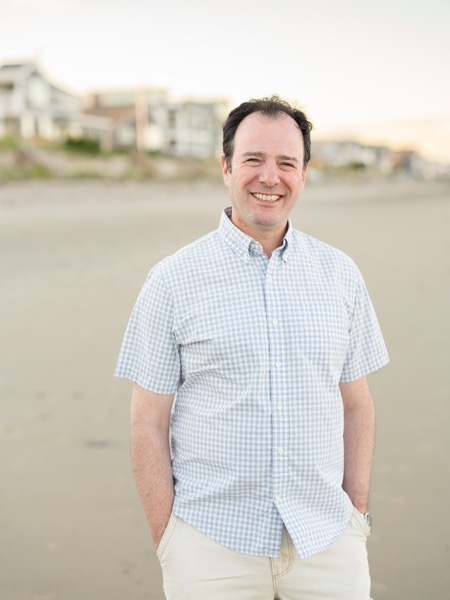 Greg Raiz on a beach at dusk, smiling, in a light blue checked shirt